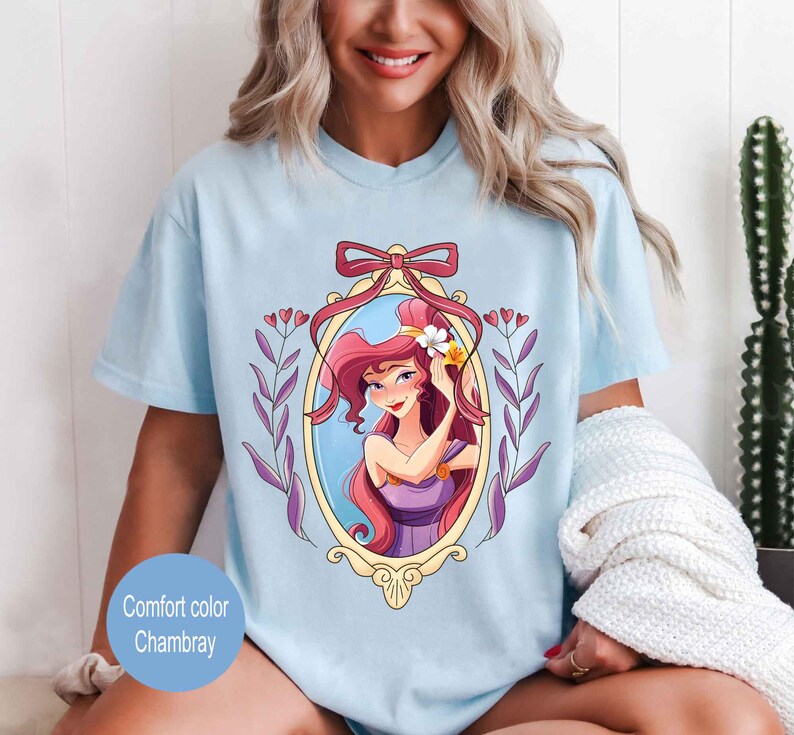 May include: A light blue t-shirt with a graphic of a cartoon woman with long red hair in a gold frame. The woman is wearing a purple dress and has a flower in her hair. The graphic is surrounded by purple flowers and leaves.