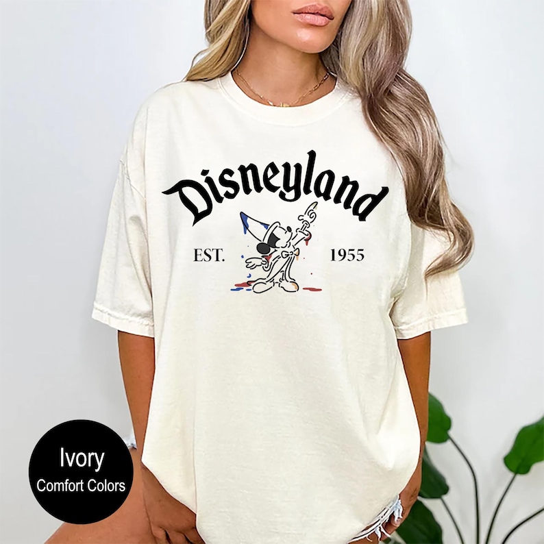 May include: A white t-shirt with a black graphic of Mickey Mouse in front of a paint splatter. The text "Disneyland" is above the graphic and "EST. 1955" is below the graphic.