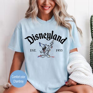 May include: Light blue t-shirt with a black graphic of Mickey Mouse in a sorcerer hat, with the text "Disneyland" in a black script font, "EST." and "1955" in black text.
