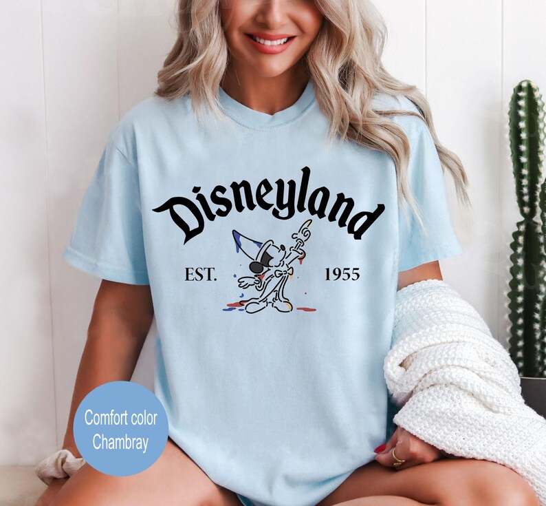 May include: Light blue t-shirt with a black graphic of Mickey Mouse in a sorcerer hat, with the text "Disneyland" in a black script font, "EST." and "1955" in black text.