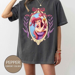 May include: A dark gray oversized t-shirt with a graphic print of a cartoon woman with long red hair in a purple dress. The woman is holding a flower in her hair. The graphic is framed in a gold oval with a purple ribbon.