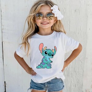 May include: A white t-shirt featuring a blue and green cartoon character with large ears wearing a blue headband. The character is standing with its arms by its side. The character is on a white background.