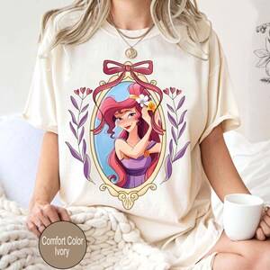 May include: A white t-shirt with a cartoon image of a woman with long red hair wearing a purple dress. The woman is framed in a gold oval with a pink bow at the top. The image is surrounded by purple and green leaves.