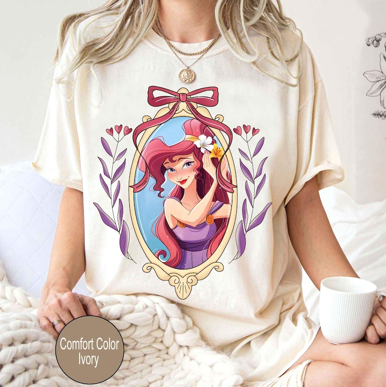 May include: A white t-shirt with a cartoon image of a woman with long red hair wearing a purple dress. The woman is framed in a gold oval with a pink bow at the top. The image is surrounded by purple and green leaves.