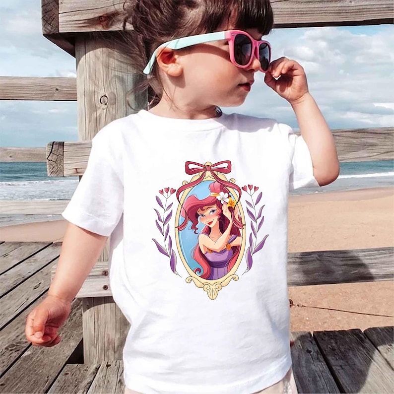 May include: White t-shirt with a cartoon image of Ariel from The Little Mermaid in a gold frame with purple flowers and vines. The image is on a white t-shirt with a bow at the top of the frame.
