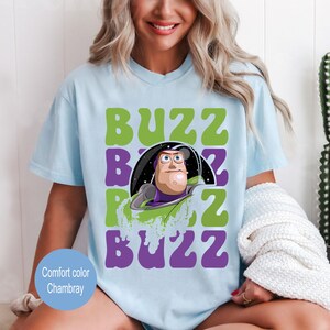 May include: Light blue t-shirt featuring a graphic of Buzz Lightyear from Toy Story. The design includes the word "BUZZ" in green and purple, with Buzz's image in the center. The shirt is a comfort color chambray.