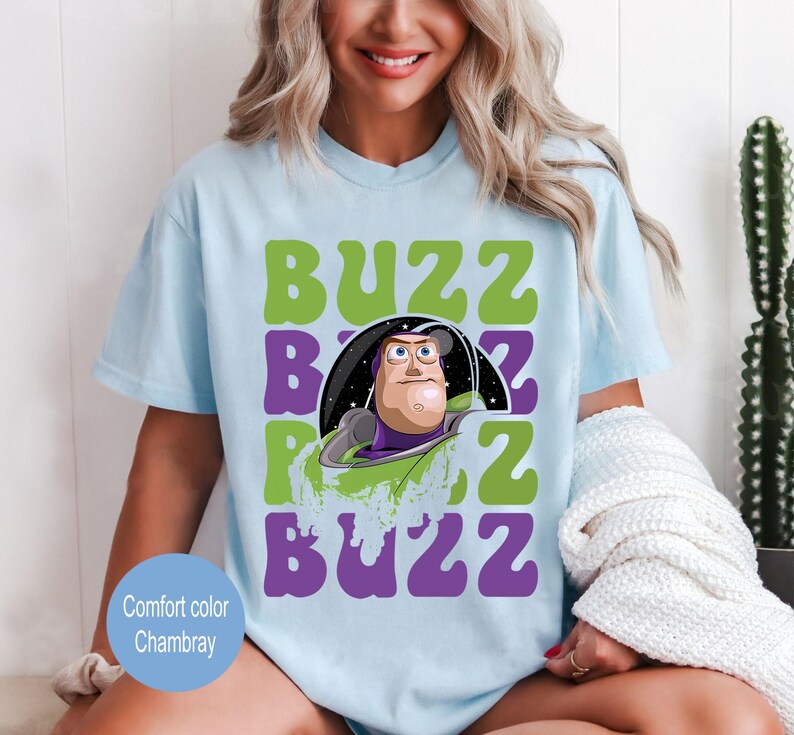 May include: Light blue t-shirt featuring a graphic of Buzz Lightyear from Toy Story. The design includes the word "BUZZ" in green and purple, with Buzz's image in the center. The shirt is a comfort color chambray.