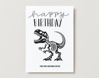 Puede incluir: Una tarjeta de felicitación blanca con texto negro y una ilustración de un esqueleto de dinosaurio. La tarjeta dice "happy BIRTHDAY" en una fuente de escritura, con "YOU PRE-HISTORIC BITCH" en la parte inferior.