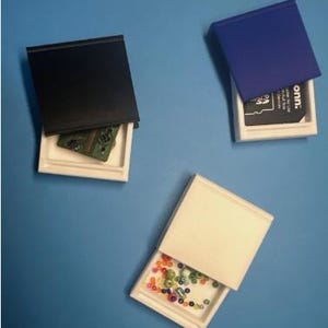 Puede incluir: Tres pequeñas cajas rectangulares blancas con tapas. Una tiene una tapa negra y una placa de circuito, otra una tapa azul y una tarjeta negra, y la tercera una tapa blanquecina y cuentas de colores.