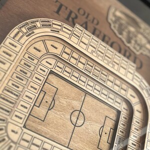 May include: A wooden model of Old Trafford, the home stadium of Manchester United Football Club. The model shows the seating layout of the stadium, including the pitch and the stands. The model is made of wood and has a brown finish.
