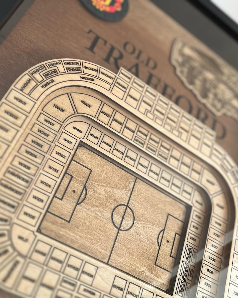 May include: A wooden model of Old Trafford, the home stadium of Manchester United Football Club. The model shows the seating layout of the stadium, including the pitch and the stands. The model is made of wood and has a brown finish.