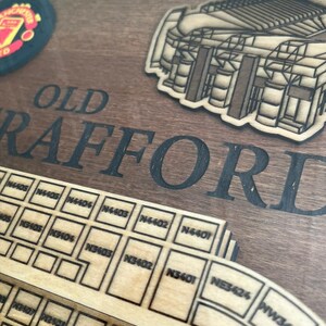 May include: A wooden sign with the words "OLD TRAFFORD" in black lettering. The sign features a laser-cut design of the stadium with seating sections labeled with numbers. The sign is a great way to show your support for Manchester United.