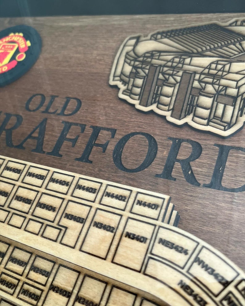 May include: A wooden sign with the words "OLD TRAFFORD" in black lettering. The sign features a laser-cut design of the stadium with seating sections labeled with numbers. The sign is a great way to show your support for Manchester United.