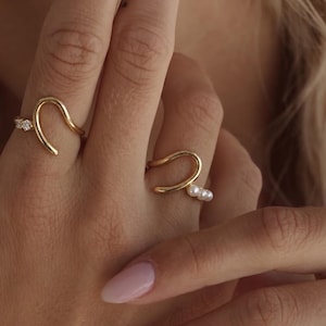 May include: Two gold-toned open rings worn on adjacent fingers. One ring features a smooth, curved gold band accented with three small clear stones. The second ring displays a similar curved gold band adorned with three small white pearls.