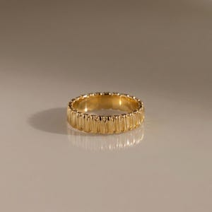 May include: A gold band ring featuring a continuous series of vertical, slightly rounded ridges around the entire exterior surface. The metal has a polished, reflective finish that catches the light, creating a uniform, textured appearance.