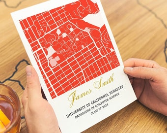 May include: A personalized map print with a red city grid design. The print includes the name "James Smith", university details, and "Class of 2024" in gold script. The print is held over a wooden table with a drink.