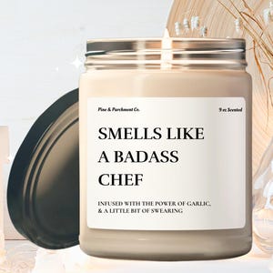 May include: A 9 oz scented candle in a clear glass jar with a black lid. The candle is labeled "Smells Like A Badass Chef" and says "Infused with the power of garlic, & a little bit of swearing."