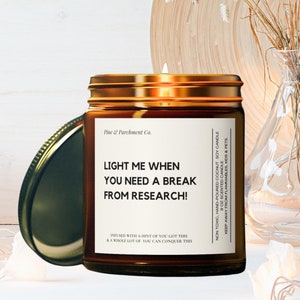 May include: A brown glass candle jar with a gold lid and a white label that reads "Light me when you need a break from research!" The candle is surrounded by white lights and a wooden tray.