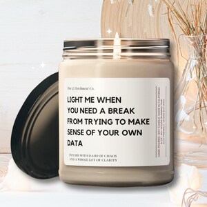 May include: A beige candle in a glass jar with a silver lid, featuring the text "LIGHT ME WHEN YOU NEED A BREAK FROM TRYING TO MAKE SENSE OF YOUR OWN DATA." The candle is next to a black lid, decorative vases, and a wooden plate.