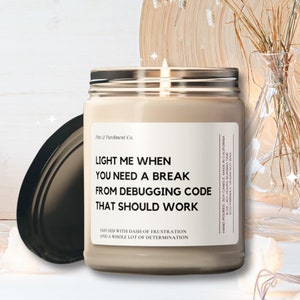 May include: A lit, beige-colored candle in a glass jar with a silver lid. The label reads "LIGHT ME WHEN YOU NEED A BREAK FROM DEBUGGING CODE THAT SHOULD WORK." The candle is next to a wooden plate, glass vases, and decorative lights.