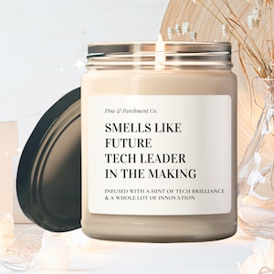 May include: A beige candle with a black label that reads "Smells Like Future Tech Leader In The Making. Infused with a hint of tech brilliance & a whole lot of innovation."