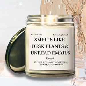 May include: A glass jar candle with a silver lid, labeled "Smells Like Desk Plants & Unread Emails." The candle is 9 oz and made with coconut soy wax. The label also reads "Infused with ambition, success & endless possibilities."