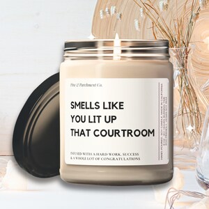 May include: A lit candle in a glass jar with a silver lid, labeled "Smells Like You Lit Up That Courtroom." The candle sits near a decorative wooden plate, a small vase with dried flowers, and a string of fairy lights. The candle's label also says "Infused with a hard work, success & a whole lot of congratulations."