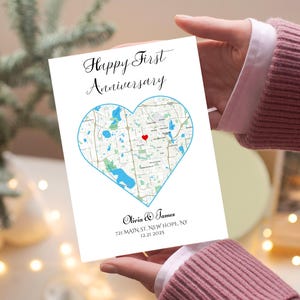 May include: A white card with the text "Happy First Anniversary" and a heart-shaped map design. The map is outlined in blue and features a red heart marker. The names "Olivia & James" and the date are also printed on the card.
