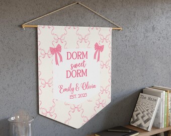 May include: A decorative banner with a cream background and pink bow pattern. The banner features the text "DORM sweet DORM" and the names "Emily & Olivia EST. 2023" in pink. The banner is hanging on a wooden rod with a natural twine hanger.