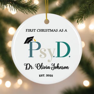 May include: White ceramic Christmas ornament with the text "First Christmas as a Psy.D. Dr. Olivia Johnson Est. 2025". A graduation cap is above the text "Psy.D."