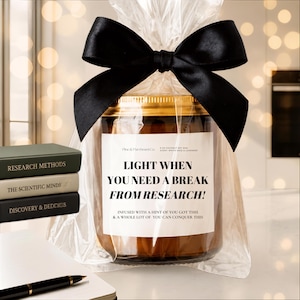 May include: A glass candle jar with a gold lid, wrapped in clear plastic with a large black satin bow. The label reads: Pine & Parchment Co. 8 oz coconut soy wax, white sage & lavender, LIGHT WHEN YOU NEED A BREAK FROM RESEARCH! INFUSED WITH A HINT OF YOU GOT THIS & A WHOLE LOT OF YOU CAN CONQUER THIS.