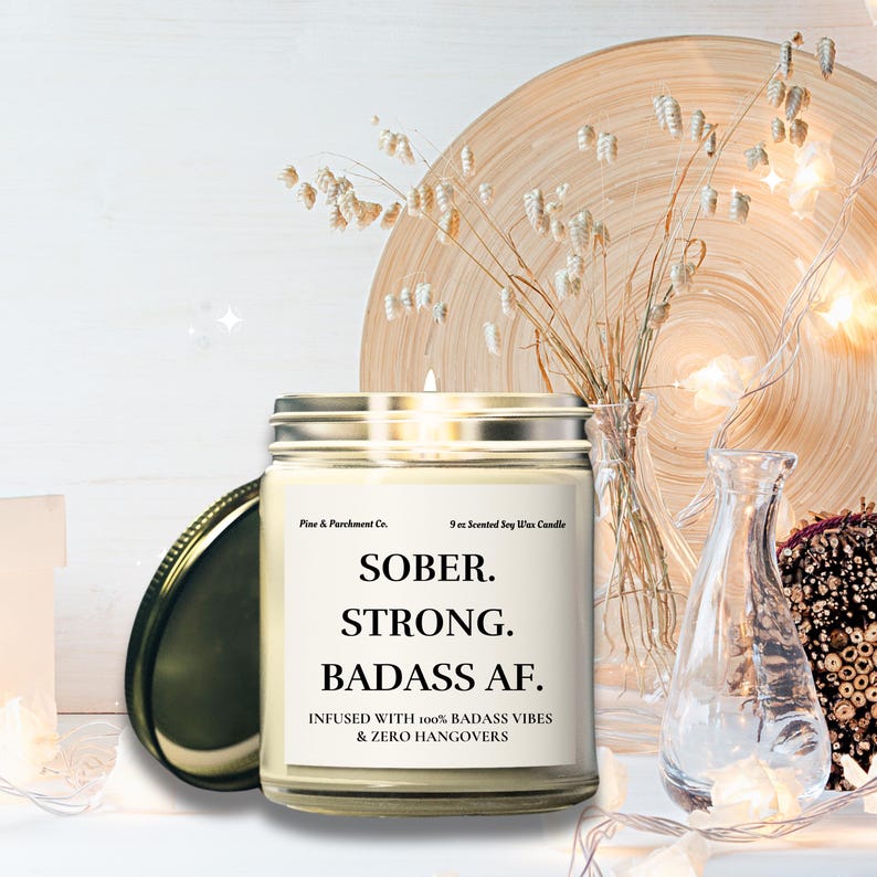 May include: A glass jar candle with a silver lid, featuring the text "SOBER. STRONG. BADASS AF." The candle is labeled "INFUSED WITH 100% BADASS VIBES & ZERO HANGOVERS." The candle is a 9 oz scented soy wax candle.