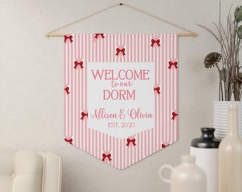 May include: A pink and white striped dorm room banner with red bows. The banner reads "WELCOME to our DORM" with the names "Allison & Olivia" and the year "EST. 2025". The banner is hanging from a wooden rod.
