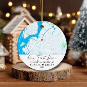 May include: Round ceramic ornament featuring a map design with a blue water section and a red pin. The text "Our First Home" is displayed, along with coordinates, names, and the year 2027. The ornament hangs from a gold string.