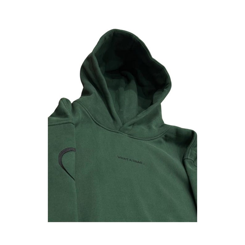May include: A dark green hoodie with a hood and long sleeves. The hoodie has a black embroidered heart on the left sleeve and the words "WHAT A DRAG" printed in black on the front.