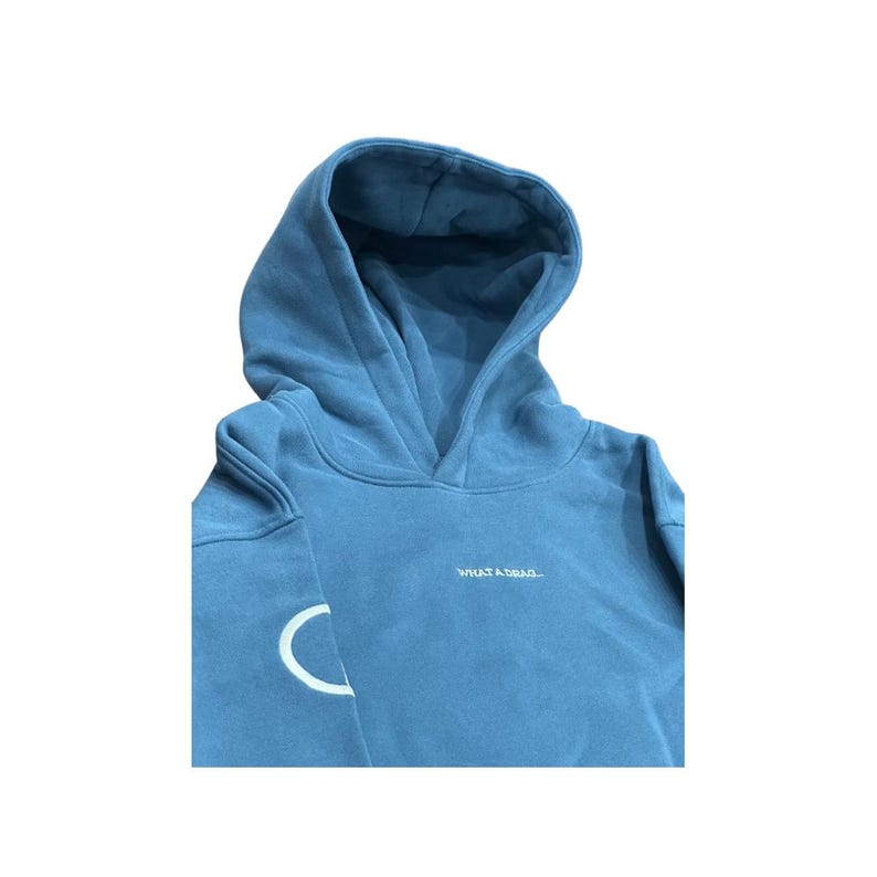 May include: A blue hoodie with a hood and long sleeves. The text "WHAT A DRAG..." is embroidered in white on the chest. A white crescent moon design is on the left sleeve.