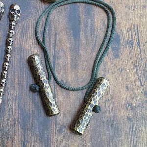 Puede incluir: Un collar con dos cilindros de latón martillado y un cordón verde oscuro. Los cilindros tienen una superficie texturizada y están conectados por un cordón anudado. Otros objetos metálicos son visibles en el fondo.