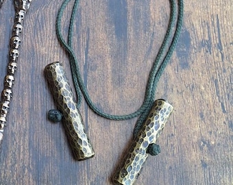 May include: A necklace featuring two hammered brass cylinders with a dark green cord. The cylinders have a textured surface and are connected by a knotted cord. Other metal objects are visible in the background.