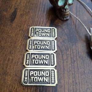 Puede incluir: Cuatro boletos color bronce de "Pound Town" con el texto "ONE-WAY" y el número "696969" impreso. Una calavera de bronce con detalles turquesa está en el fondo. Los objetos están sobre una superficie de madera oscura.