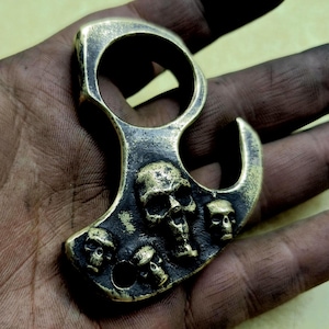 May include: Brass knuckles with a skull design. The knuckles have a ring for the finger and a hook on the side. The skulls are raised and detailed.