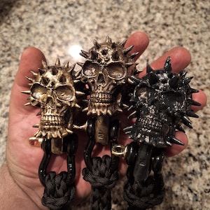 May include: Three skull-shaped keychains with spiked details. The keychains are made of metal and have a black, gold, and silver finish. Each keychain is attached to a black braided rope.