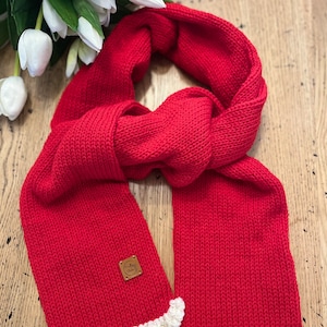 May include: A vibrant red knitted scarf with a cream-coloured ruffled edge. The scarf features a small, square leather tag. The scarf is displayed on a wooden surface with white tulips in the background.