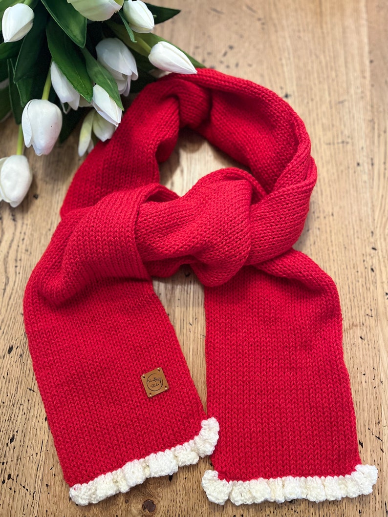 May include: A vibrant red knitted scarf with a cream-coloured ruffled edge. The scarf features a small, square leather tag. The scarf is displayed on a wooden surface with white tulips in the background.