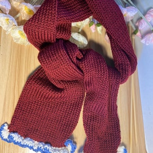 May include: A burgundy knitted scarf with a ruffled edge. The scarf is made of a thick, textured yarn and has a white and blue trim along the bottom edge. The scarf is displayed on a light wooden surface.