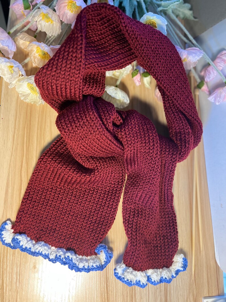 May include: A burgundy knitted scarf with a ruffled edge. The scarf is made of a thick, textured yarn and has a white and blue trim along the bottom edge. The scarf is displayed on a light wooden surface.