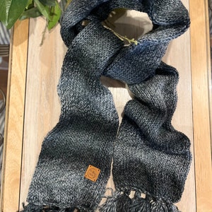 May include: A knitted scarf in grey and blue tones, with a gradient design. The scarf is rectangular with tassels at the ends and a small, square label. The scarf is made from a soft, textured yarn, perfect for colder weather.