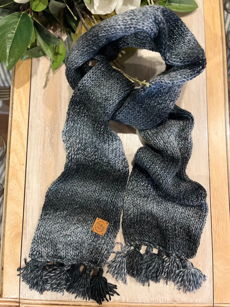 May include: A knitted scarf in grey and blue tones, with a gradient design. The scarf is rectangular with tassels at the ends and a small, square label. The scarf is made from a soft, textured yarn, perfect for colder weather.