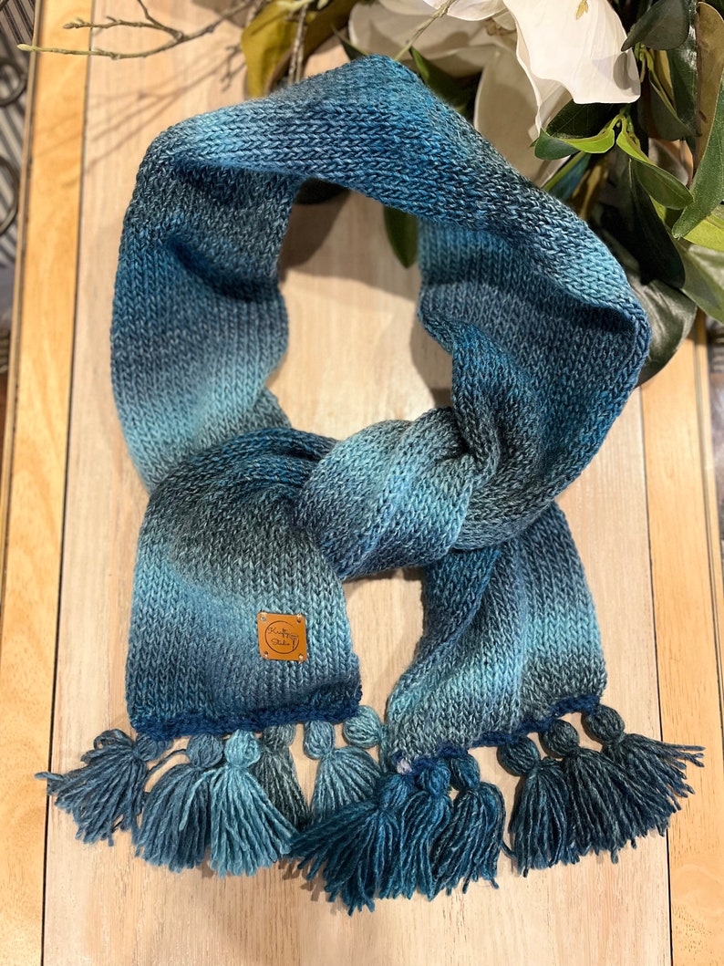 May include: A knitted scarf in shades of blue, with a gradient effect from dark to light. The scarf has a textured knit design and is finished with tassels. A small, brown leather tag is sewn on the scarf.