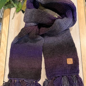May include: Hand-knitted scarf in shades of purple, grey, and black, with a gradient effect. The scarf features a rectangular shape with tassels at the ends and a small, square leather tag. The scarf is made of soft yarn.