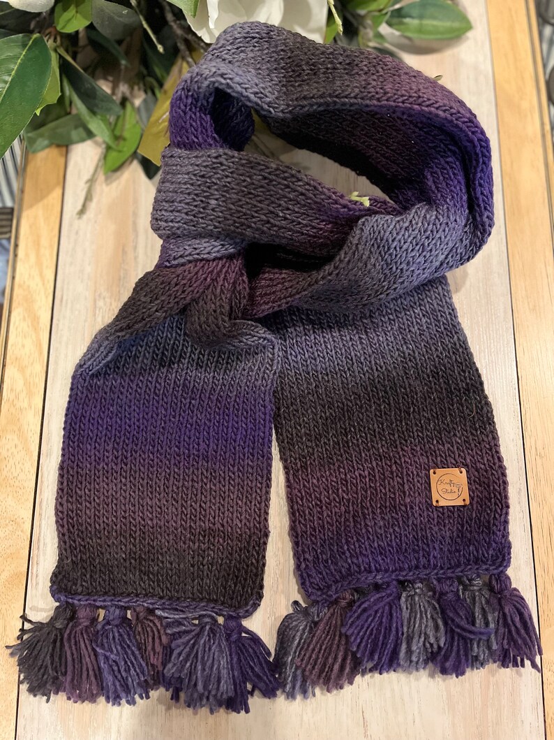 May include: Hand-knitted scarf in shades of purple, grey, and black, with a gradient effect. The scarf features a rectangular shape with tassels at the ends and a small, square leather tag. The scarf is made of soft yarn.